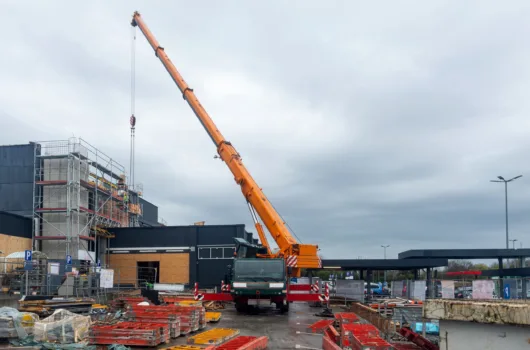 Guindaste telescópico realizando içamento em obra de construção civil com estrutura metálica e materiais no canteiro